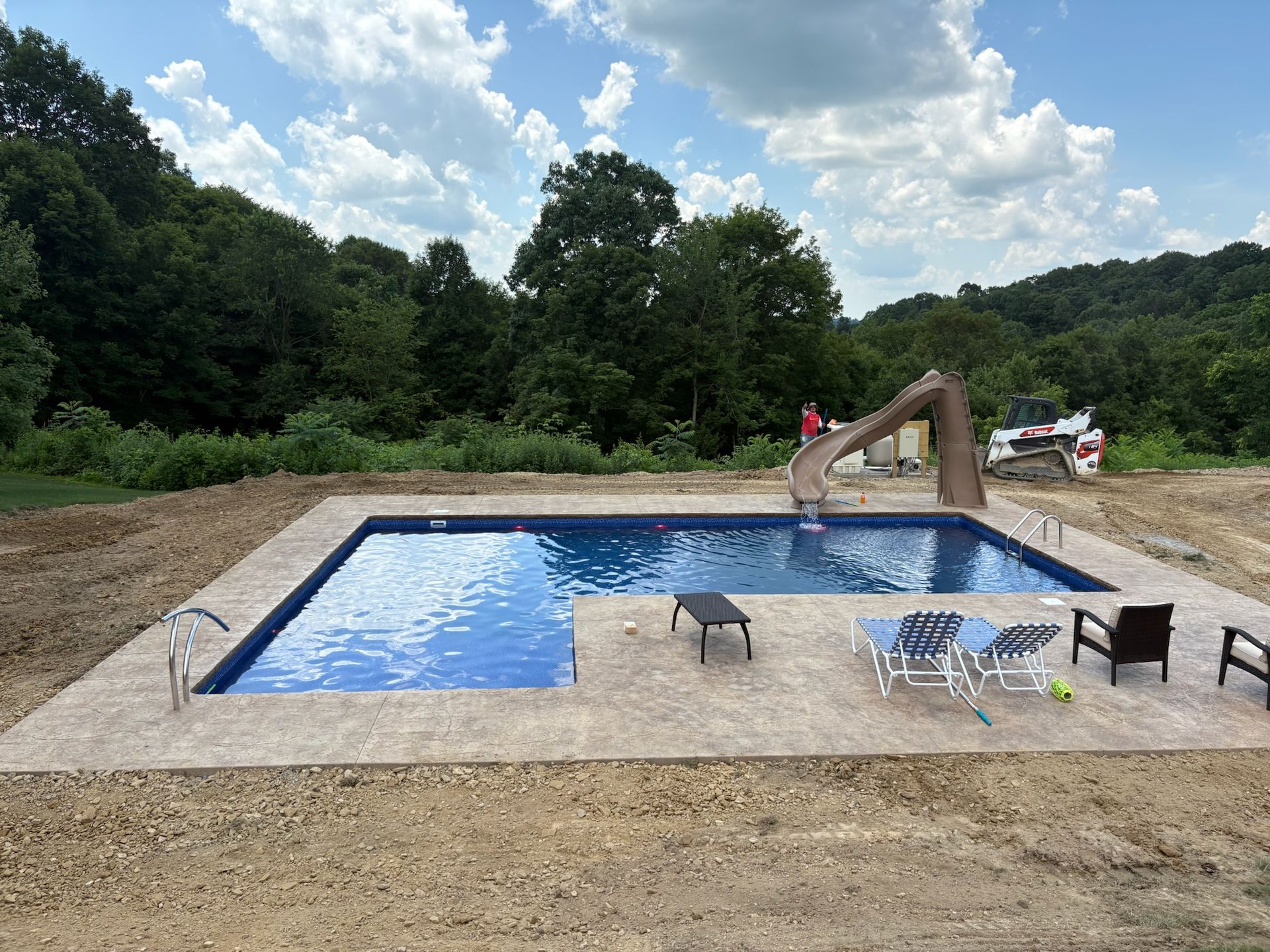 Pool with slide surrounded by concrete deck, chairs, and trees. Sunny day.