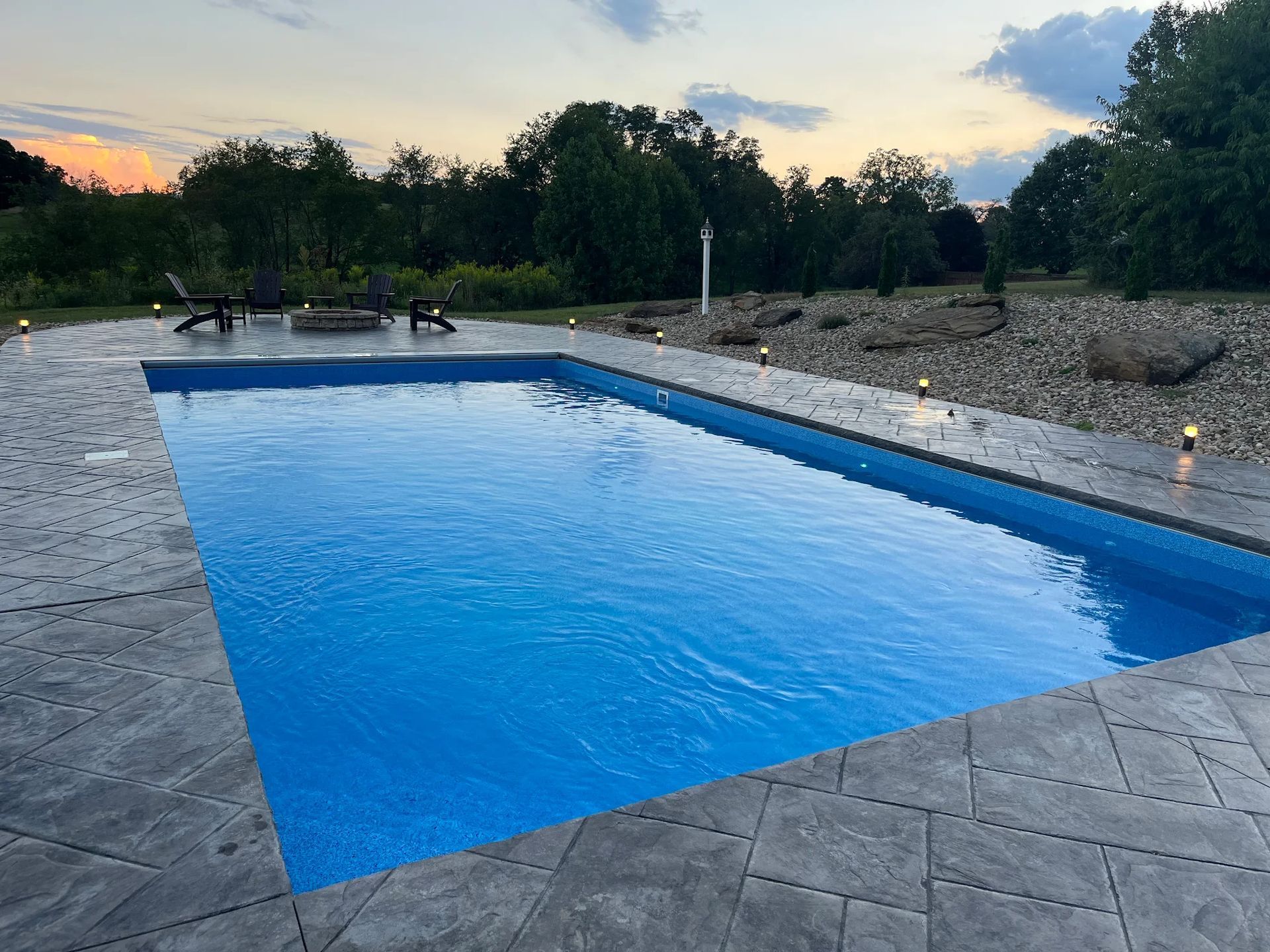 Rectangular pool with blue water and gray stone patio at dusk; trees in background.