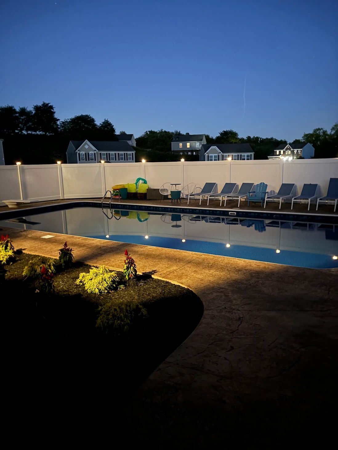 Night view of a lit pool with surrounding lights and fence. Lawn chairs are set up near the pool edge.