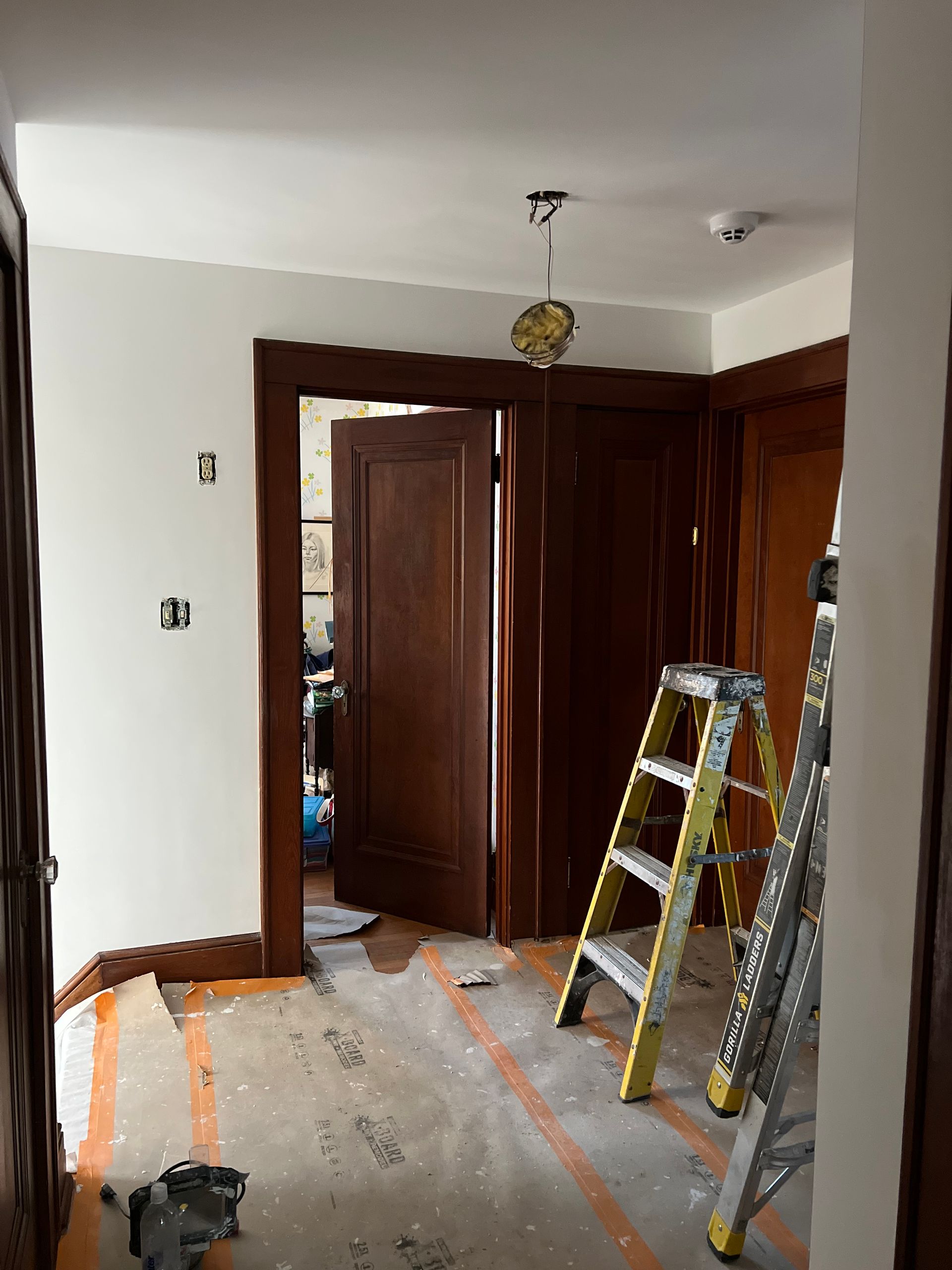 Renovation scene in a room with wooden doors, a ladder, and a bare floor with protective tape.