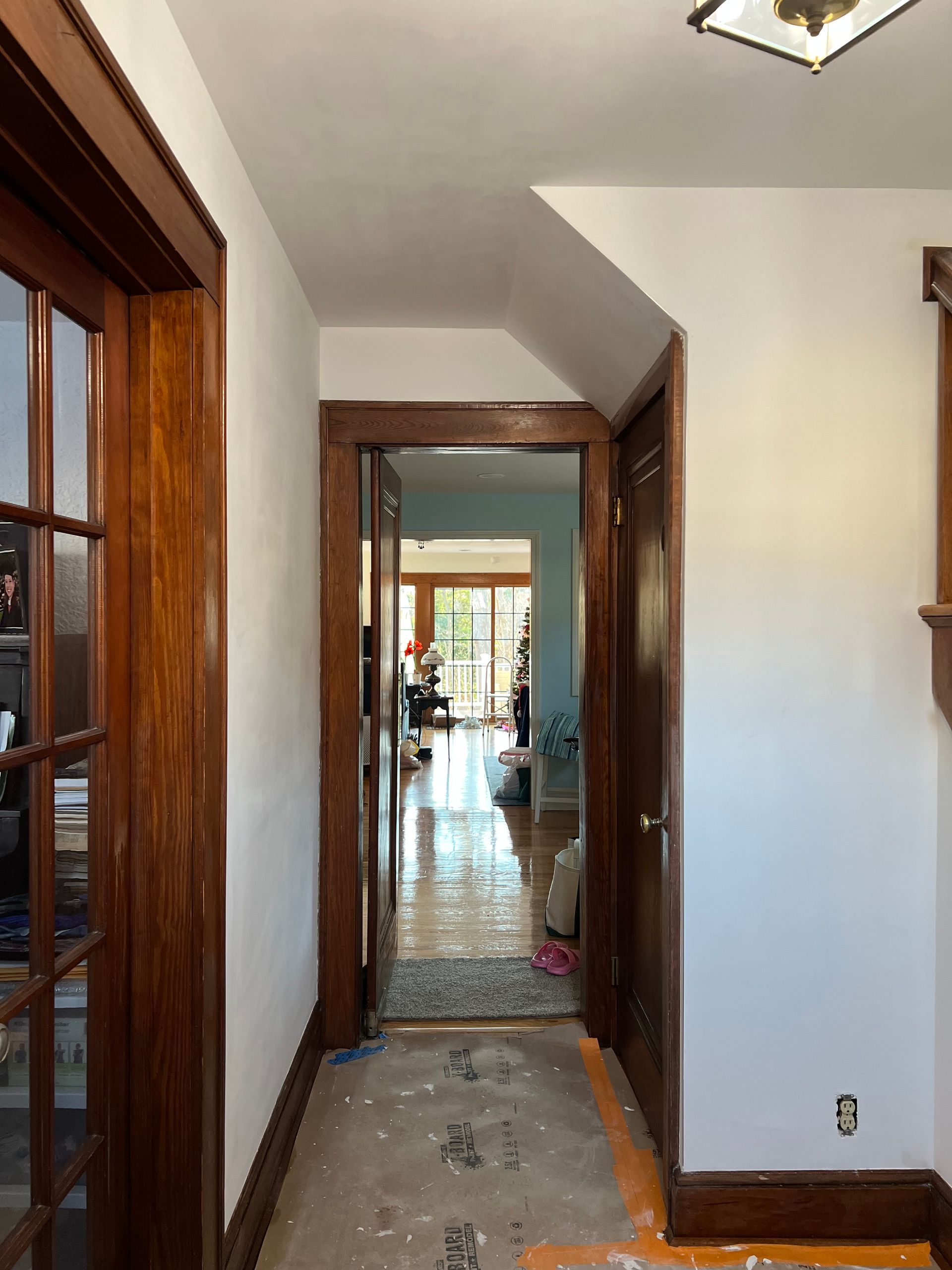 Narrow hallway with wood trim and tiled floor, leading to a bright room with windows and a distant plant.