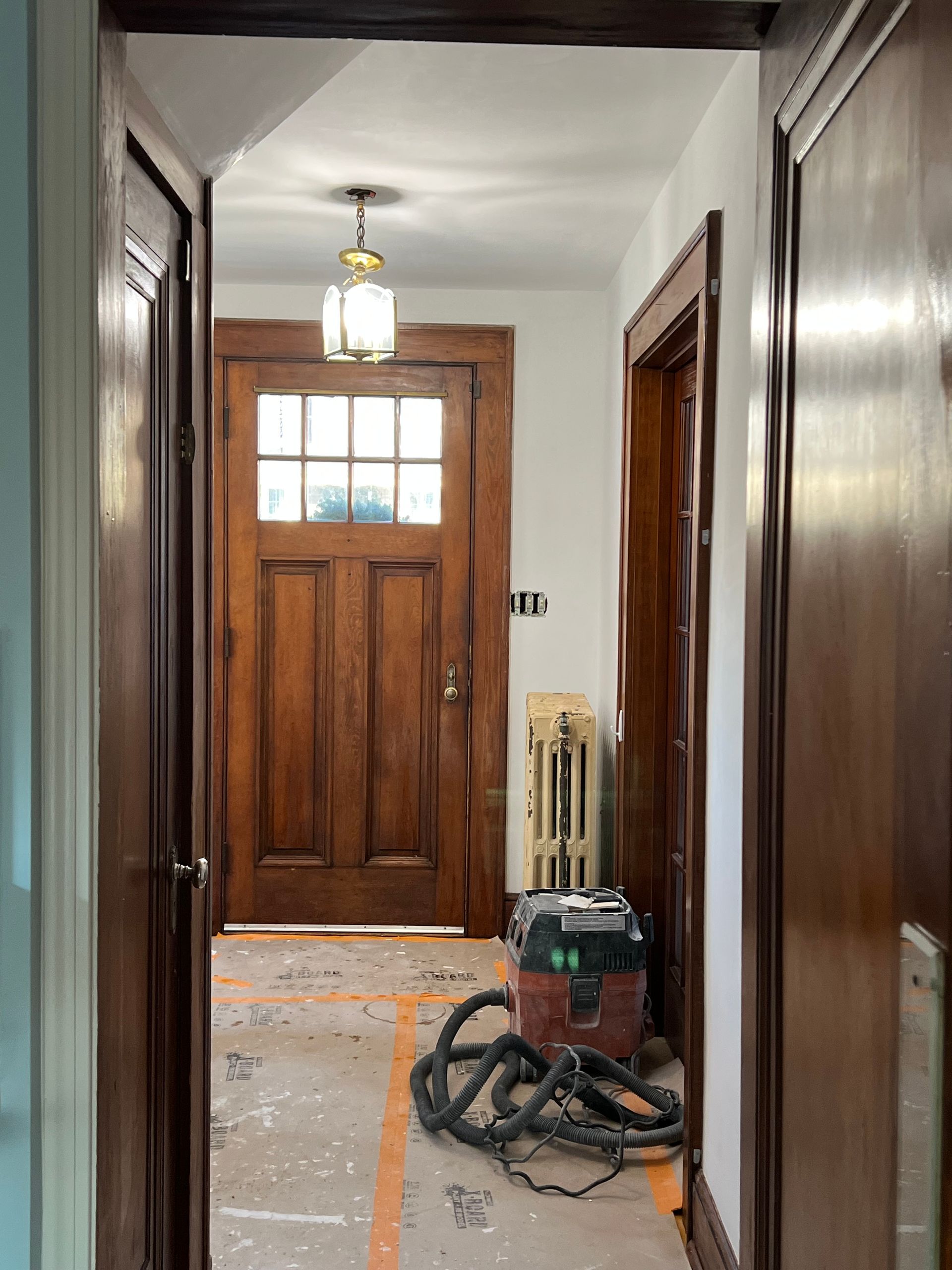 Narrow hallway with wooden front door, hallway light, and vacuum cleaner on the floor.