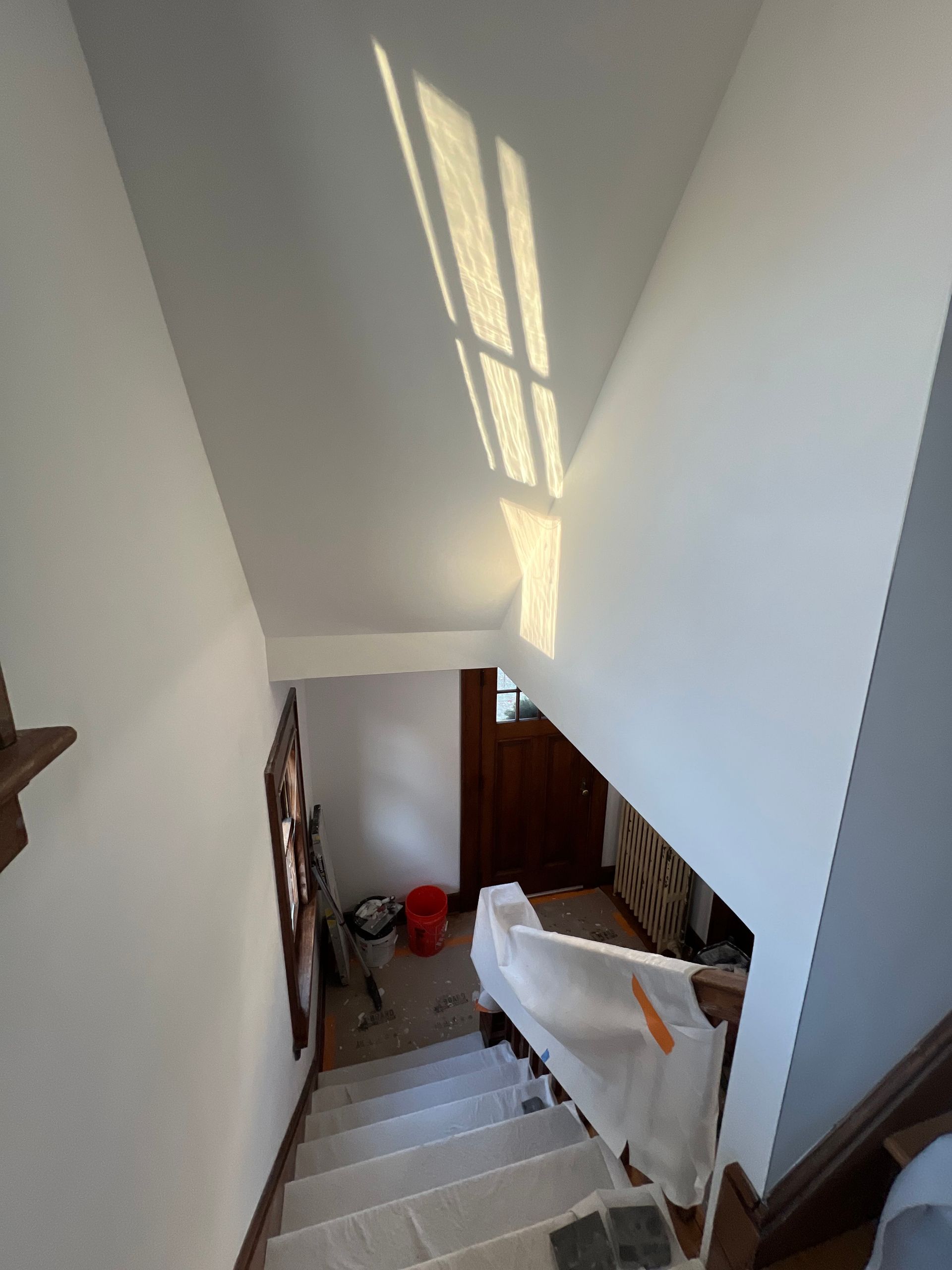Narrow staircase with white walls and sunlight, leading down to a landing with a red bucket and covered furniture
