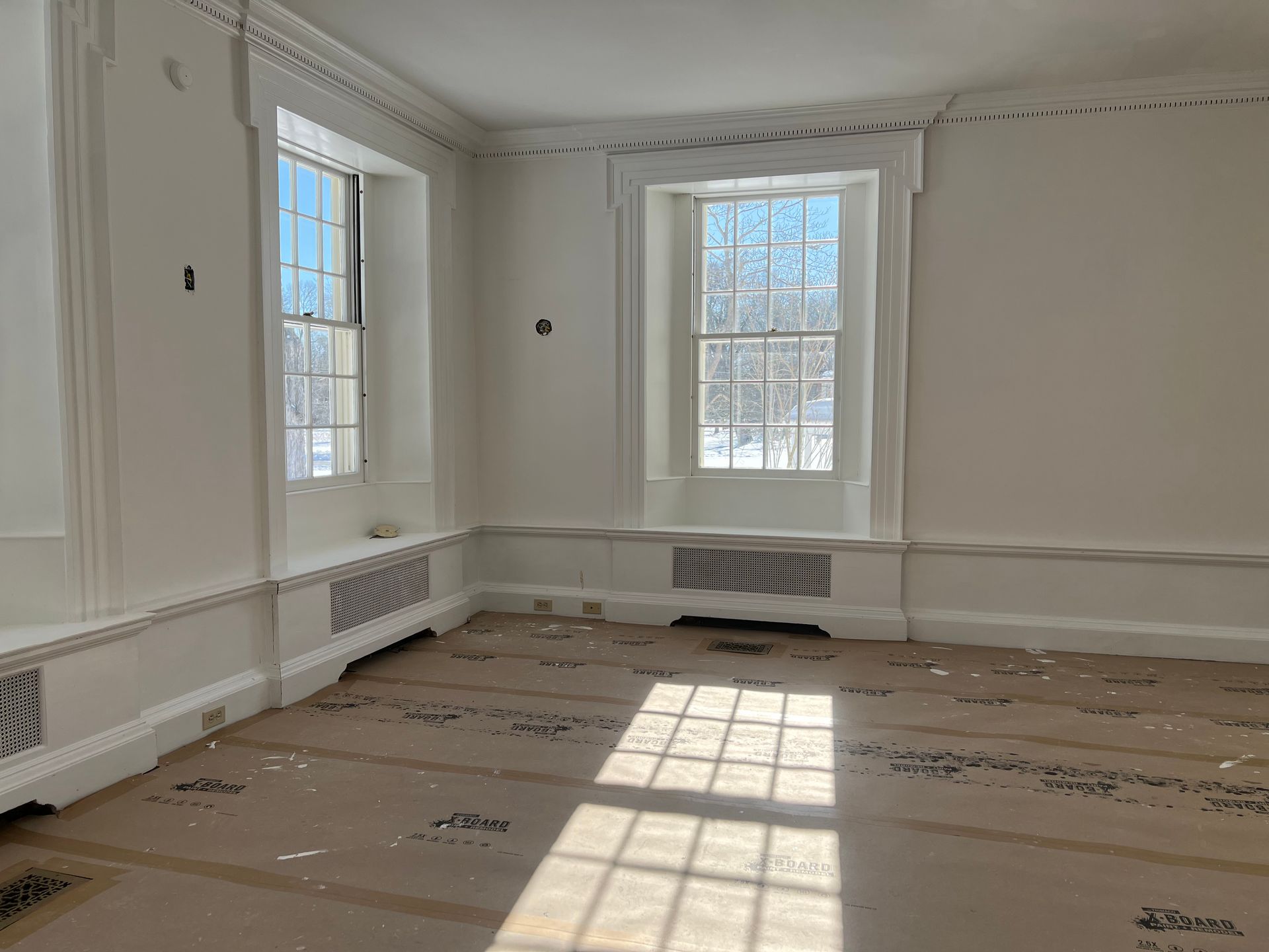 Empty white room with tall windows, sunlit floor, and unfinished baseboards.