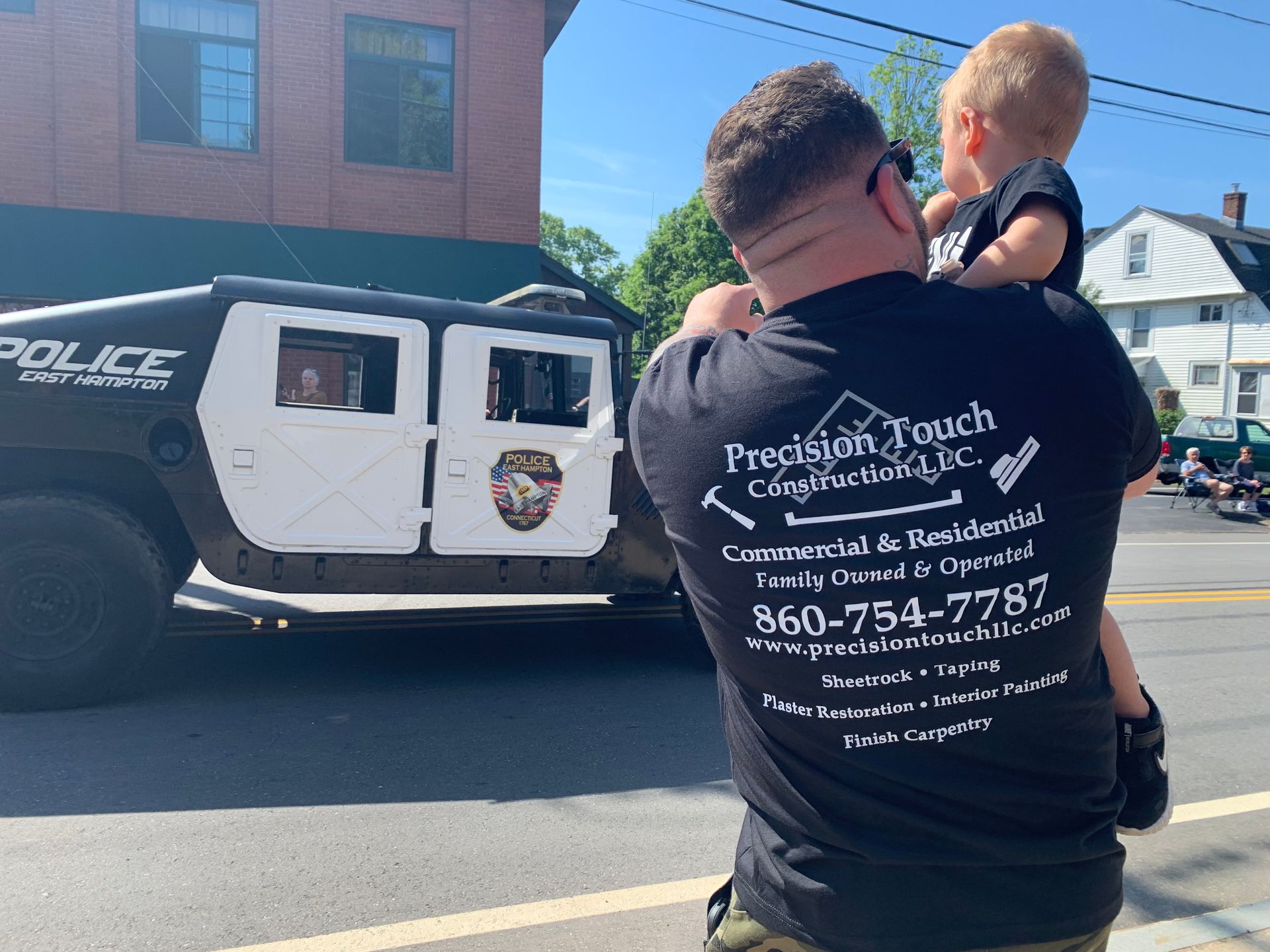 Man in black shirt carrying a child stands near a police van on a sunny street.