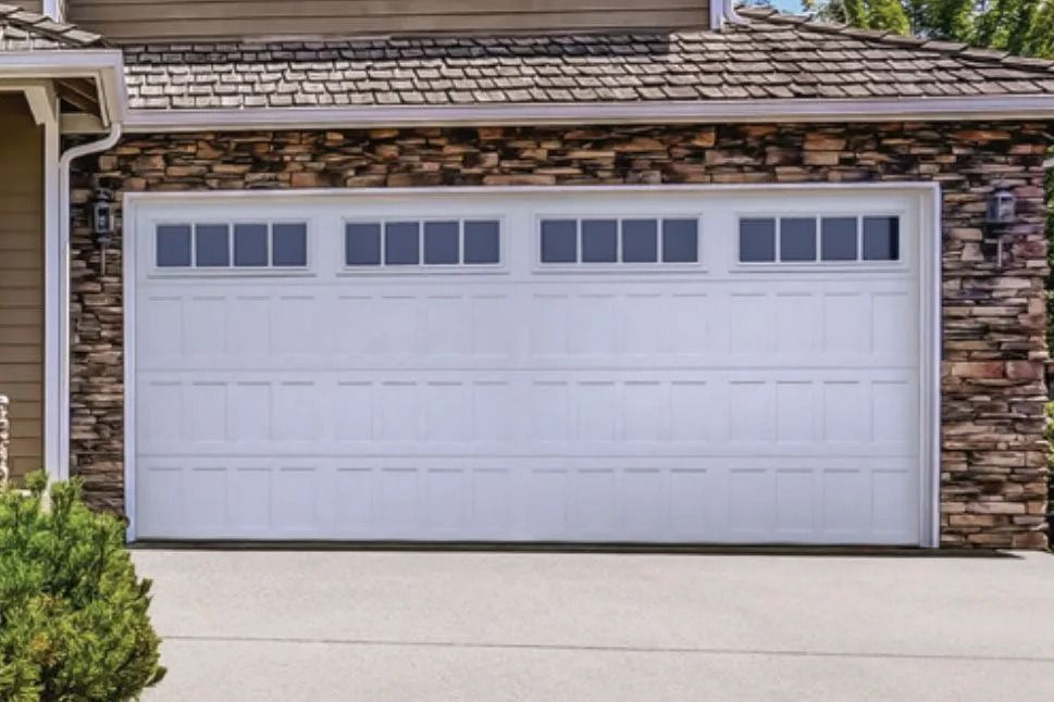 White garage door with rectangular windows above, set in a stone wall, on a concrete driveway.