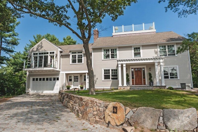Two-story light gray house with a stone wall and a driveway. It has a garage and a rooftop deck.