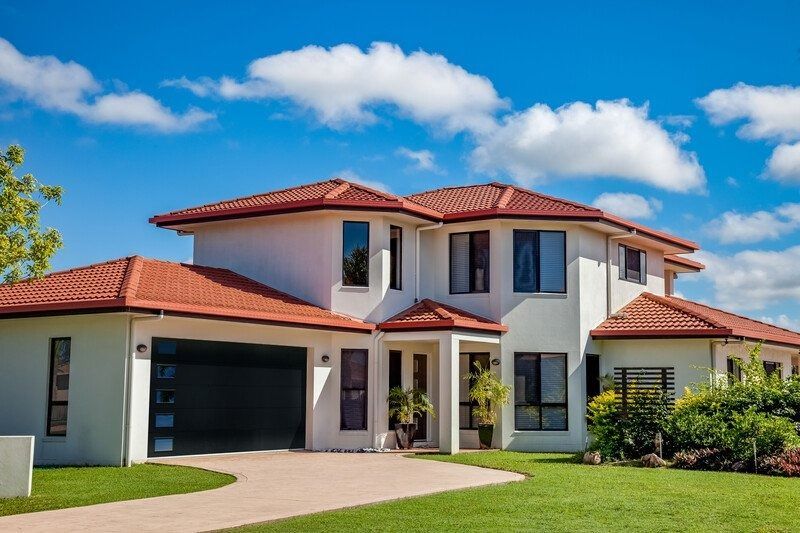White two-story house with orange tile roof, black garage door, and green lawn under a blue sky.
