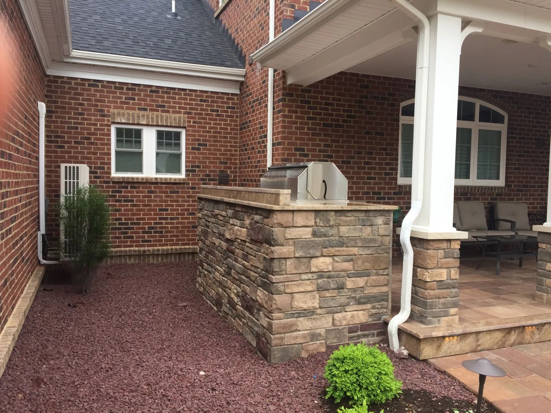 Brick house entryway with stone porch columns, red gravel path, and small green shrubs