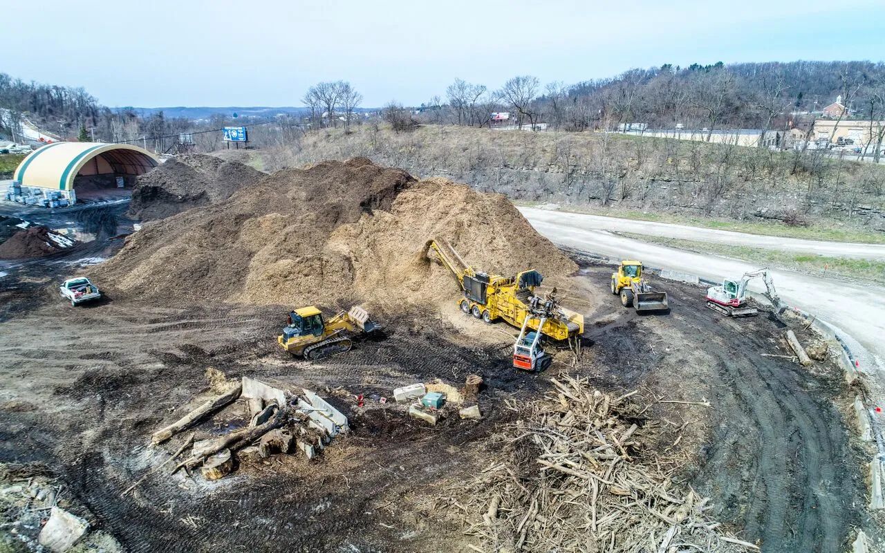 Construction site with excavators and a large dirt pile beside a road and riverbank