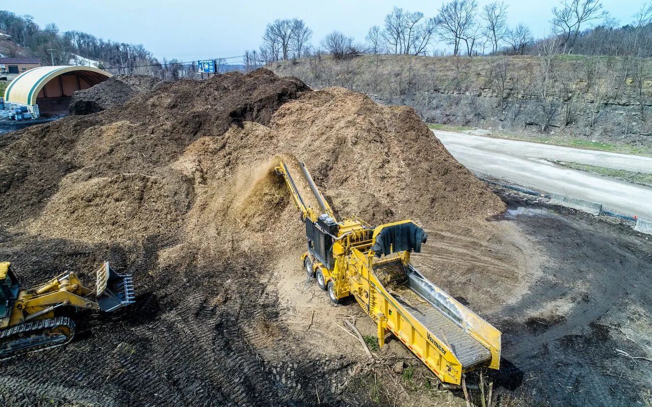 Yellow excavator on a dirt mound at a quarry beside a road, moving earth near a tunnel entrance