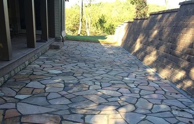 Stone walkway beside a building and retaining wall, leading toward trees in bright sunlight