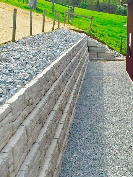 Stone retaining wall beside a gravel path on a grassy hillside, with wooden fence posts along the top.