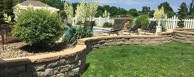 Backyard stone patio and retaining wall beside a green lawn and white fence, with shrubs and a pool in the background