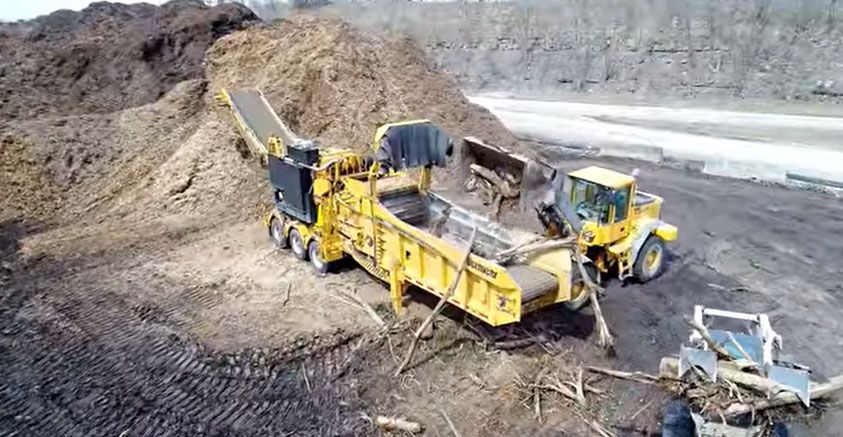 Yellow industrial machine moving soil in a dirt quarry, with a worker standing on top.