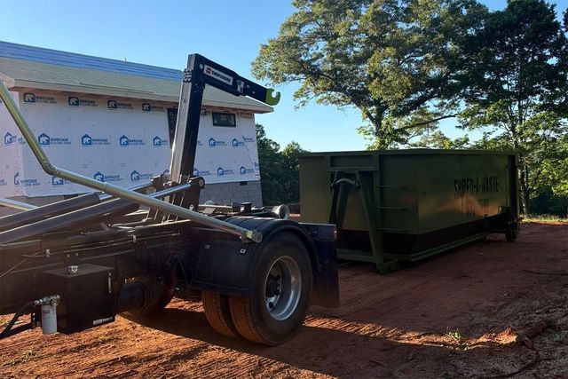 Tow truck lifting a vehicle beside a green dumpster on a sunny dirt lot