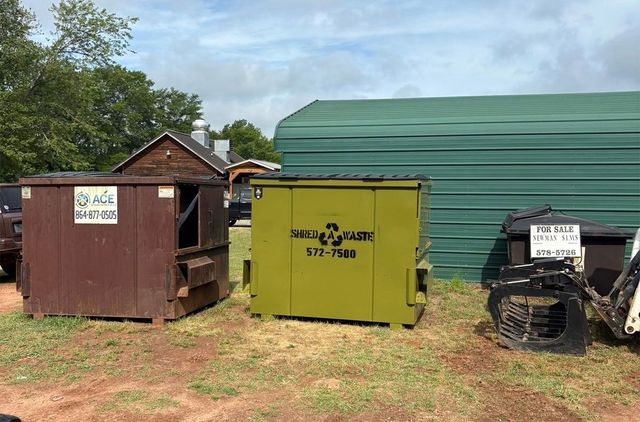 Two dumpsters and a trash bin beside a green building in a grassy lot