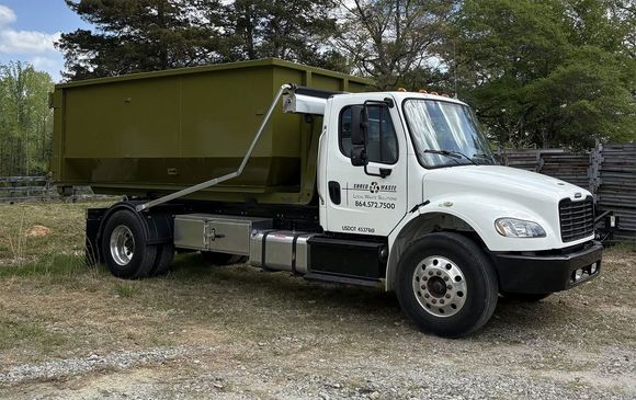 White dump truck with green container parked on a gravel lot near trees