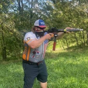 Man aiming a rifle outdoors in a grassy wooded area, wearing a cap and shooting glasses