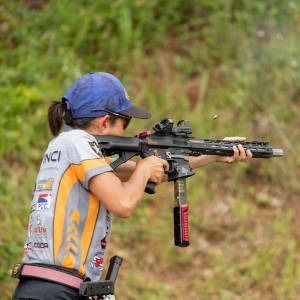Shooter aiming a rifle at an outdoor target range, wearing a blue cap and gray competition jersey.