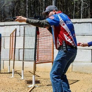 Man aiming a handgun at an outdoor shooting range, wearing a blue jersey and cap.