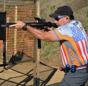 Shooter aiming a rifle at an outdoor range, wearing a cap, sunglasses, and an American flag jersey.