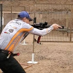 Shooter in blue cap and white jersey aiming a rifle at an outdoor range