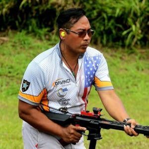 Shooter aiming a rifle outdoors, wearing safety glasses and ear protection, in a grassy field.