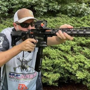 Person aiming a black rifle with a red-dot sight outdoors in front of green bushes