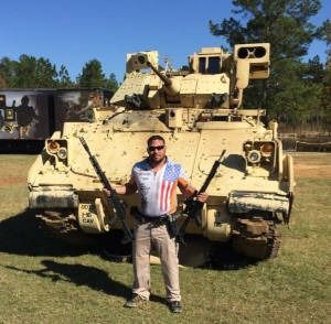 Man standing in front of a tan military vehicle on grass, holding a rifle-shaped prop
