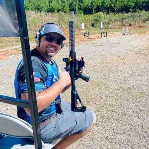 Smiling shooter seated at an outdoor range, holding a rifle with targets in the background.