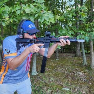 Person aiming a scoped rifle outdoors in a wooded area, wearing a blue helmet and gray shirt.