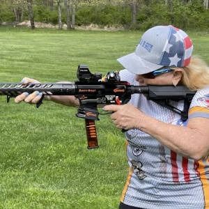 Woman aiming a black rifle on a grassy field, wearing a star-pattern cap and gray jersey.