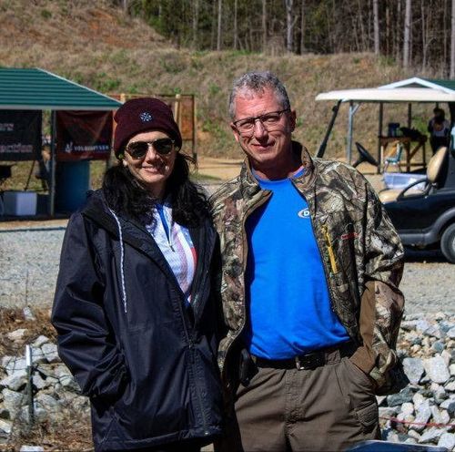 Two people in outdoor jackets and sunglasses standing together on a rocky shore.