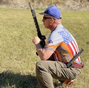 Man crouching outdoors with a rifle, wearing a blue cap and American flag shirt.