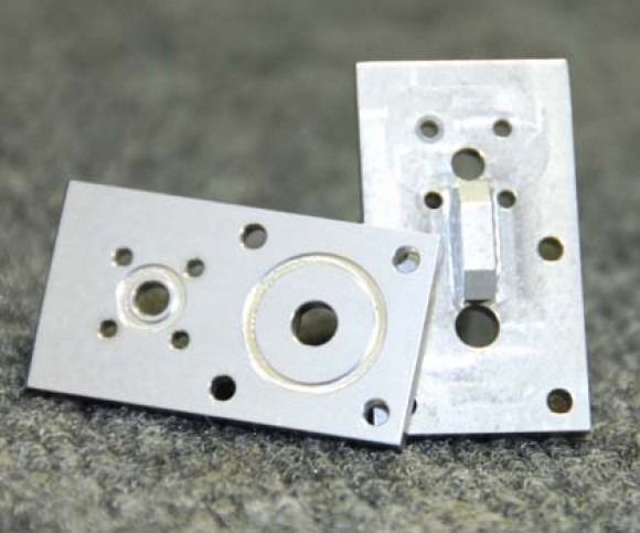 Two silver metal plates with drilled holes and circular cutouts on a gray surface