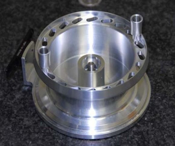 Shiny silver machined metal component with circular openings and raised posts on a dark surface