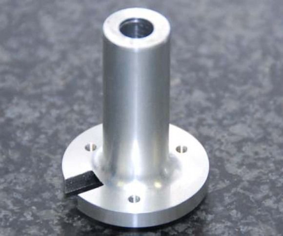 Silver metal cylindrical part with a flange, screw holes, and a black tab on a dark surface