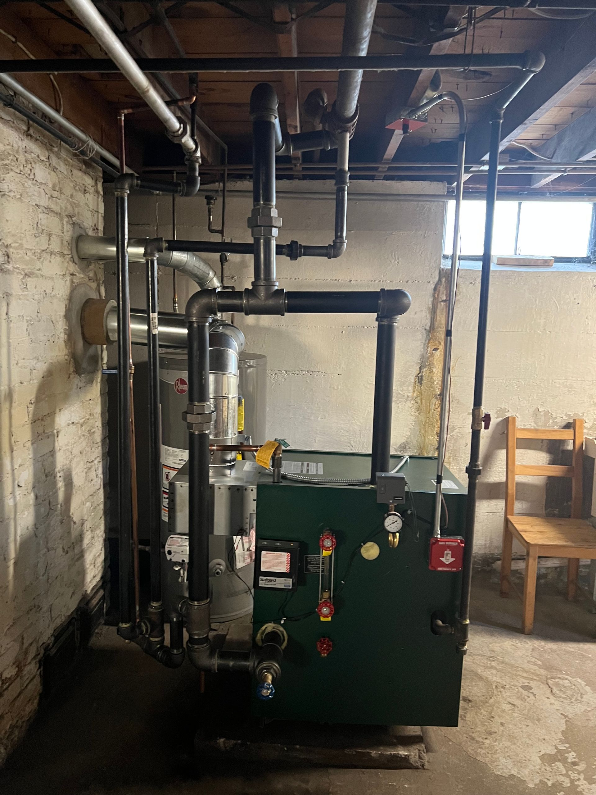 A green boiler is sitting in a basement next to a chair.