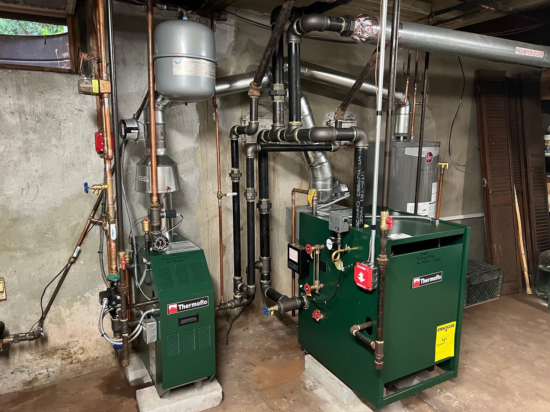 Two green boilers are sitting in a basement next to each other.