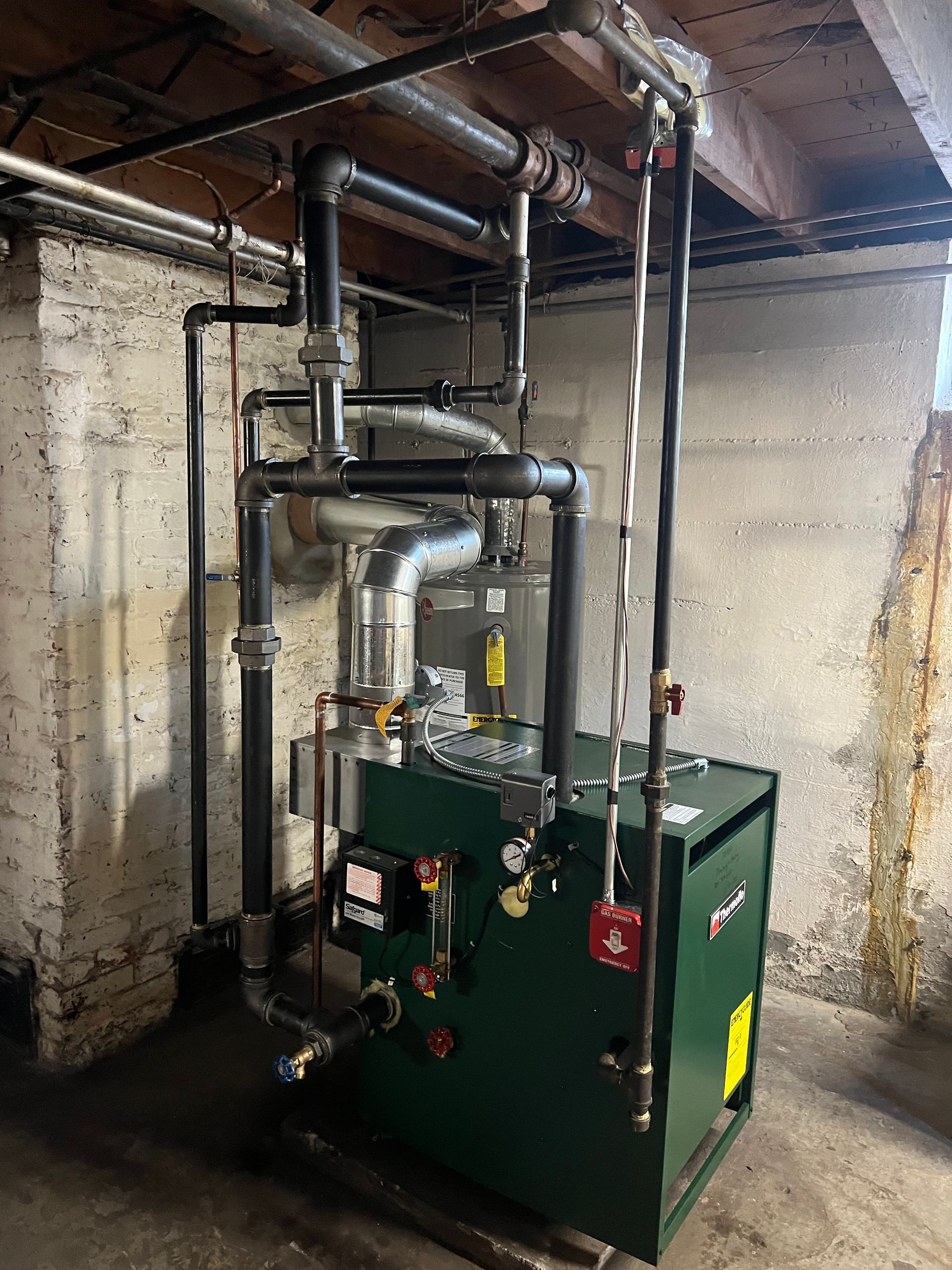 A green boiler is sitting in a basement surrounded by pipes.