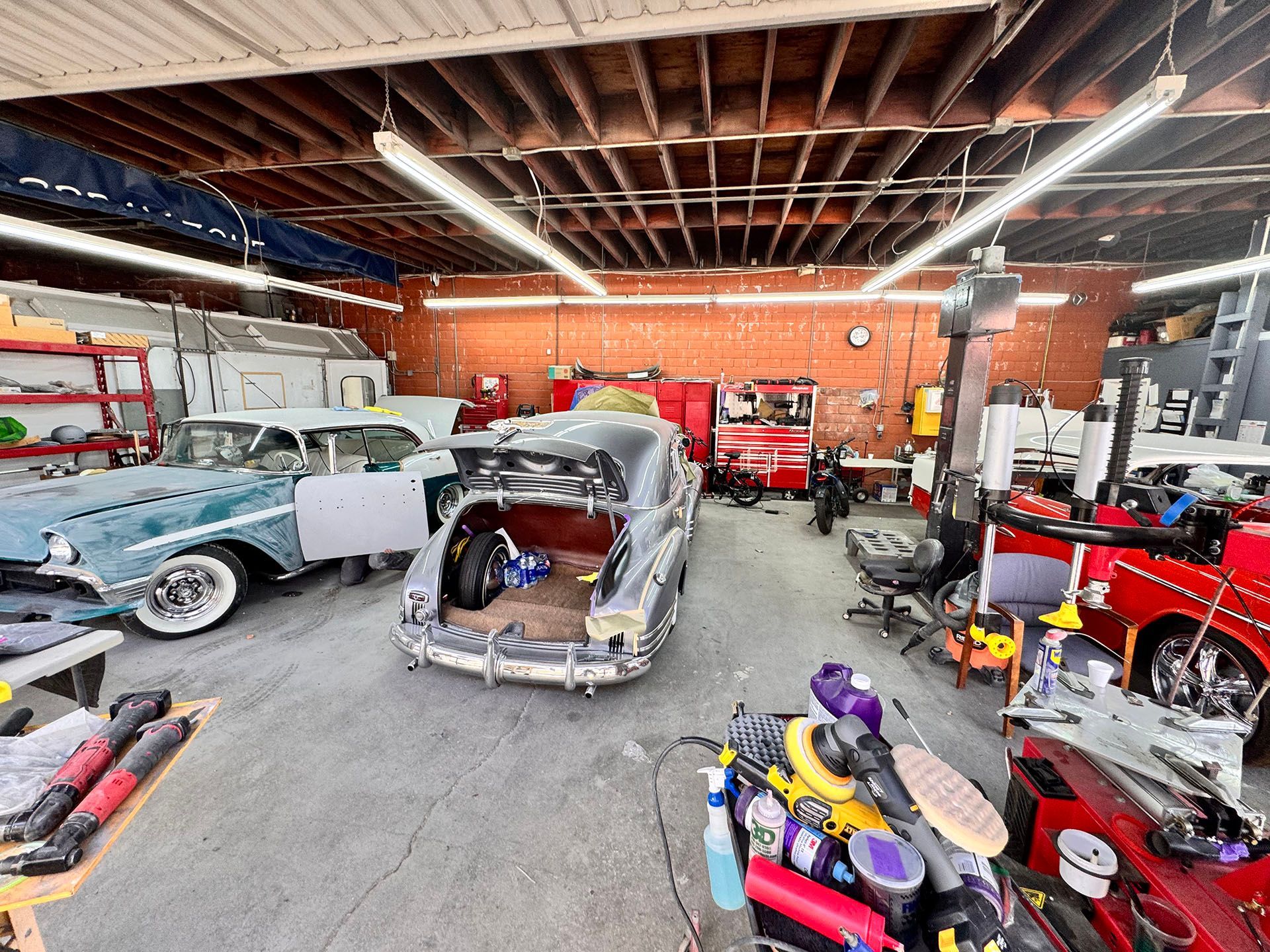 Garage with classic cars in various states of restoration. Tools and equipment are scattered. Overhead lighting.