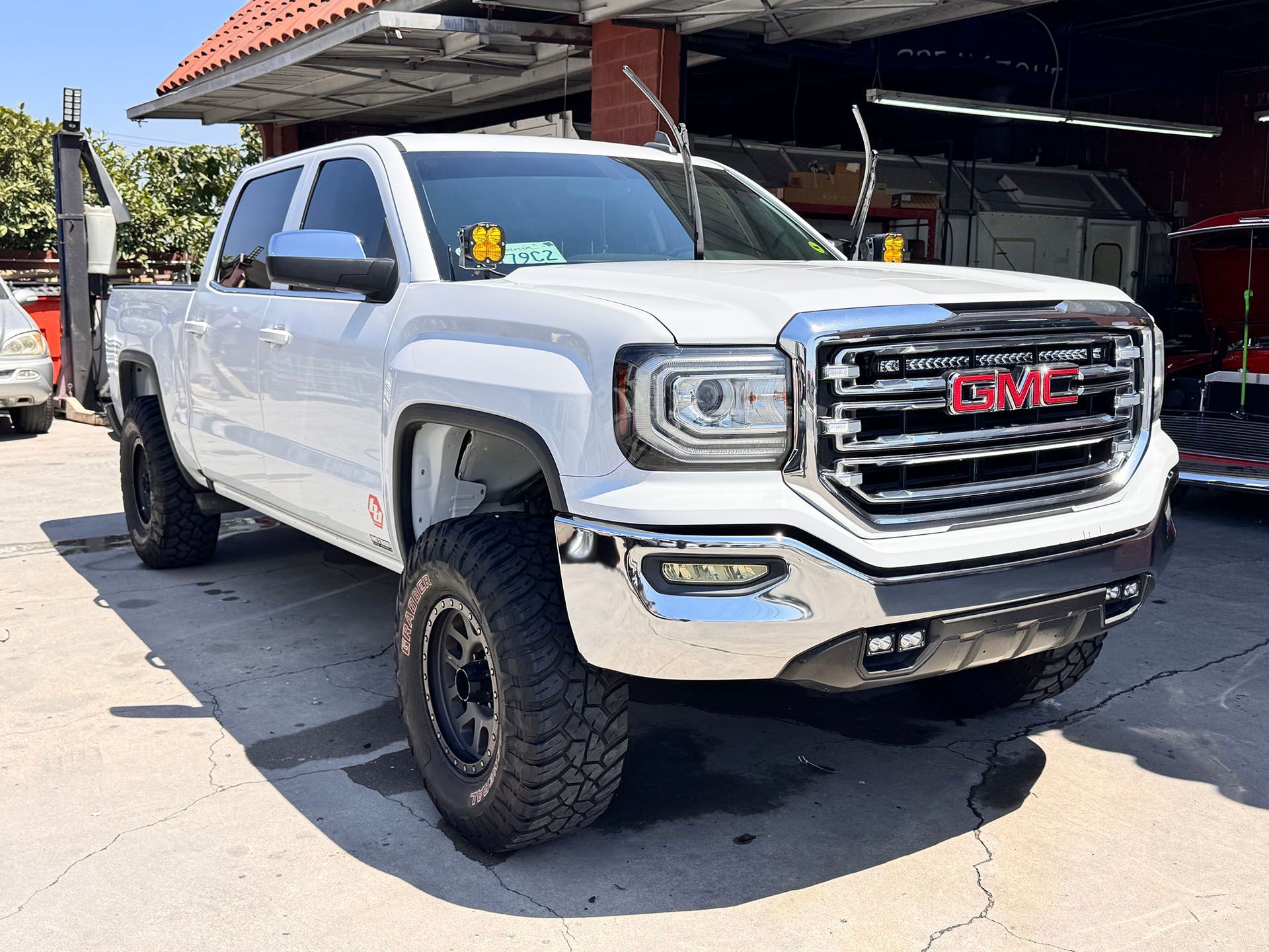 White GMC Sierra pickup truck with black off-road wheels and amber fog lights parked outside a shop.