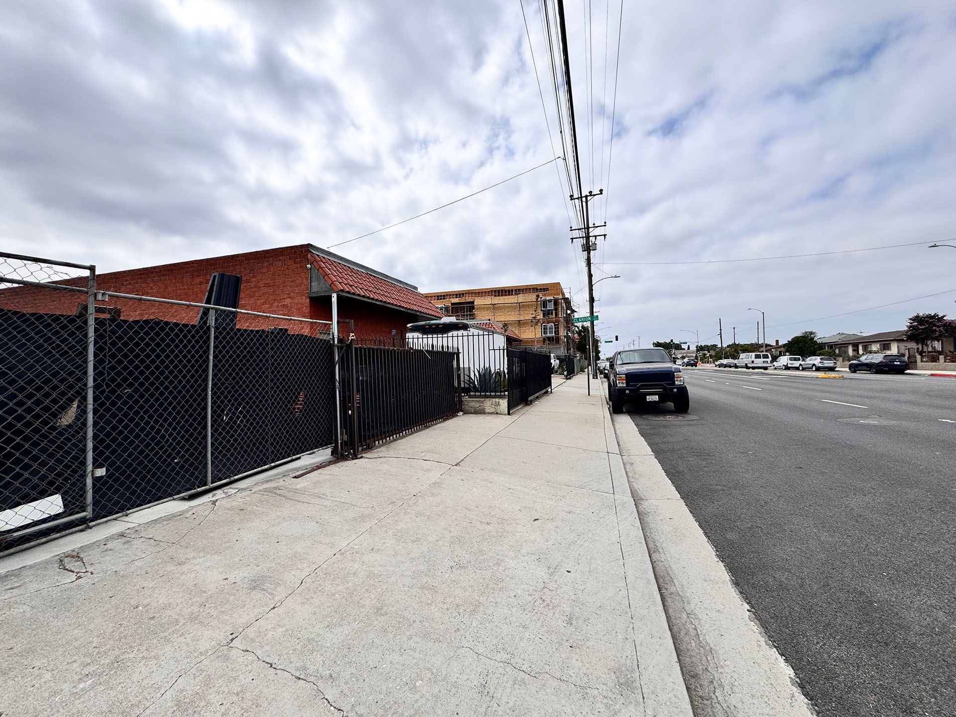 Sidewalk beside a road. Black fence and buildings on the left. Cloudy sky. Black vehicle parked on the side.