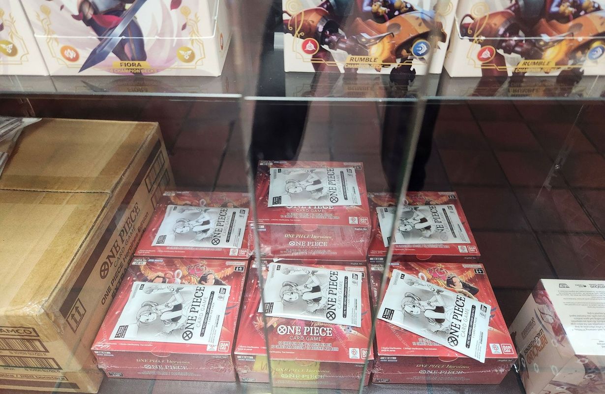 Several red boxes of trading card game booster packs are stacked on a glass store shelf.