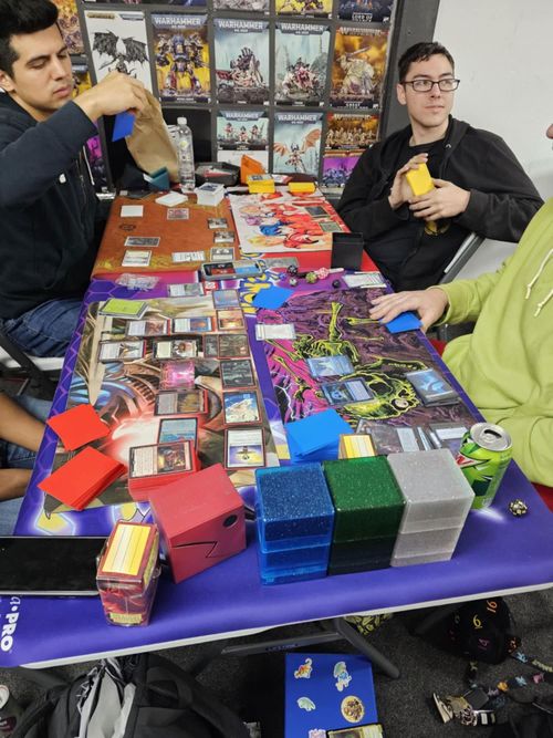 Three people playing a card game at a table filled with decks, playmats, and gaming supplies.