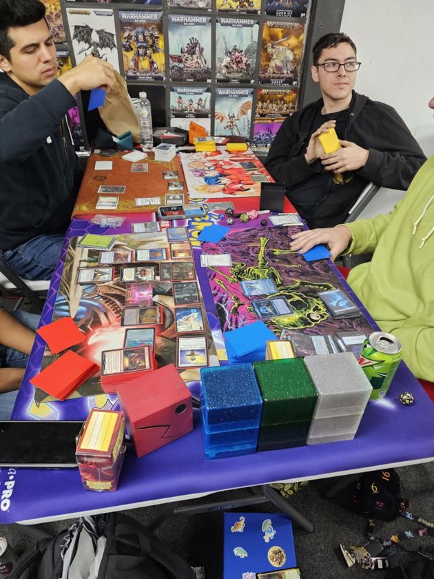 Three people playing a card game at a table filled with decks, playmats, and gaming supplies.