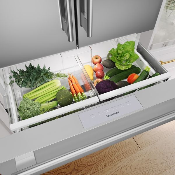 An open refrigerator drawer filled with fresh produce including leafy greens, celery, carrots, apples, and zucchini.