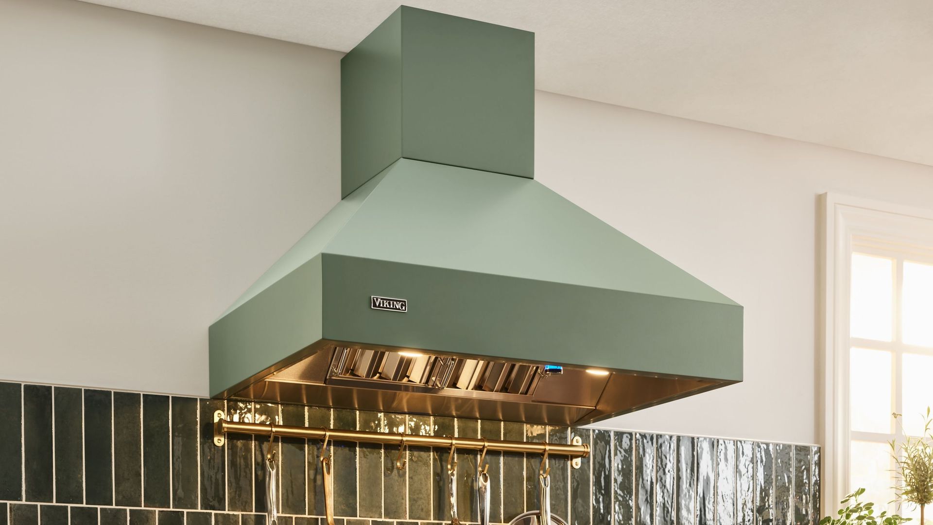 A sage green kitchen range hood is mounted above a wall with vertical, dark-colored backsplash tiles and a metal rack.