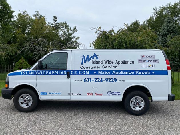 White appliance repair van with blue lettering parked outdoors near trees and gravel.